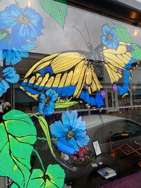 Hand‑painted window mural artwork of a colourful seasonal butterfly and flowers in a hair salon in Godalming, Surrey.