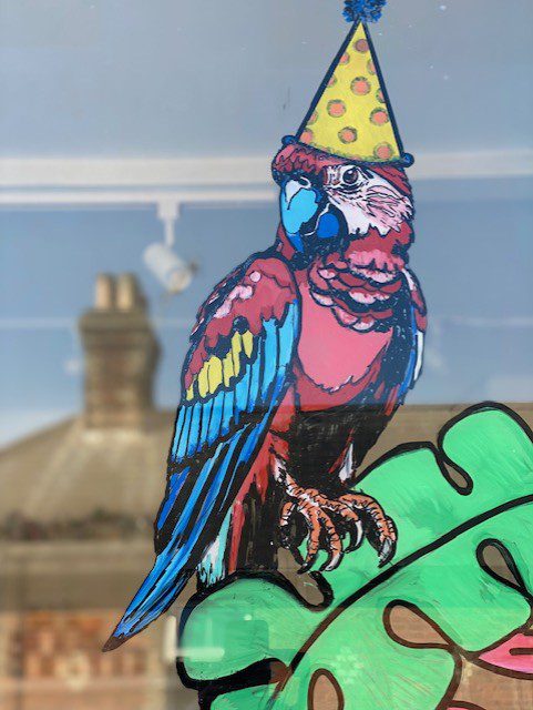 Hand‑painted window mural artwork of a colourful parrot with party hat for Party Arty craft and art room, Haslemere Surrey.