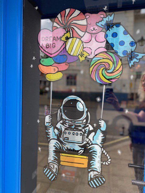 Hand‑painted window mural artwork showing an astronaut on a swing lifted by candy‑shaped balloons for the Slush Truck sweets shop in Alton Hampshire 