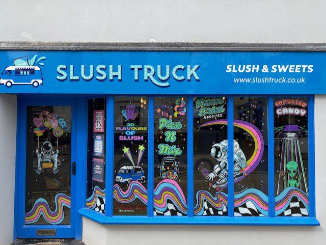 Hand‑painted window mural artwork featuring astronauts, aliens, candy graphics and rainbow waves across the Slush Truck slush and sweets shopfront in Alton Hampshire