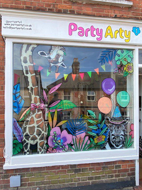 Hand‑painted window mural artwork featuring giraffe, zebra and cow with colourful balloons and tropical leaves for Party Arty craft and art room in Haslemere, Surrey.